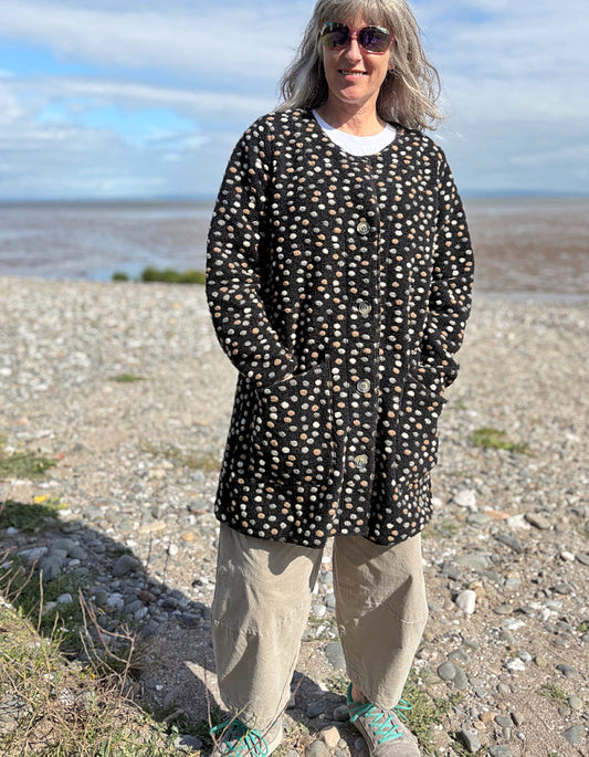 Person wearing a black polka dot coat standing on a pebbly beach with water and sky in the background.