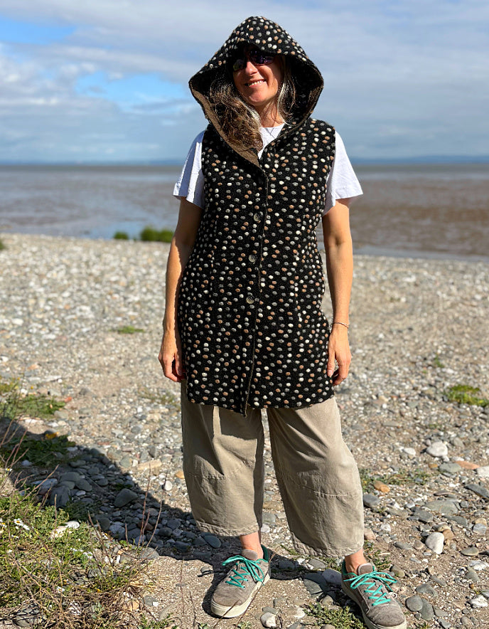 Person wearing a black patterned hooded vest and beige pants standing on a pebbly beach.