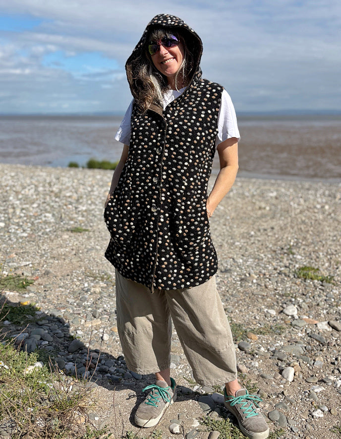 Person wearing a polka dot hooded vest standing on a pebbly beach with water in the background.