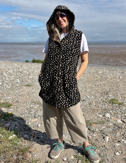 Person wearing a polka dot hooded vest standing on a pebbly beach with water in the background.