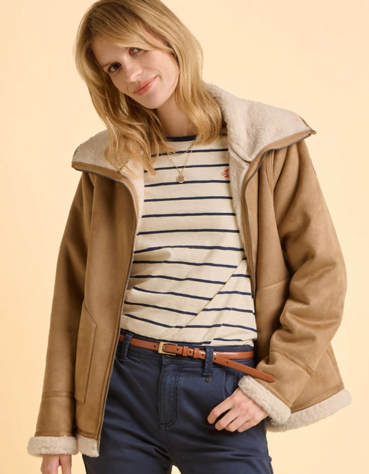 Woman wearing a brown jacket with fur lining, striped shirt, and blue jeans on a beige background