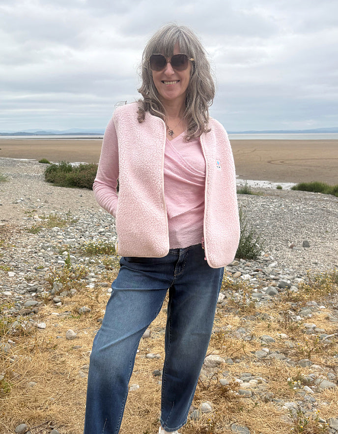 A woman wearing a light pink sherpa zip-up vest over a matching pink top, blue jeans, and sunglasses, standing outdoors on a rocky, dry beach landscape under an overcast sky.