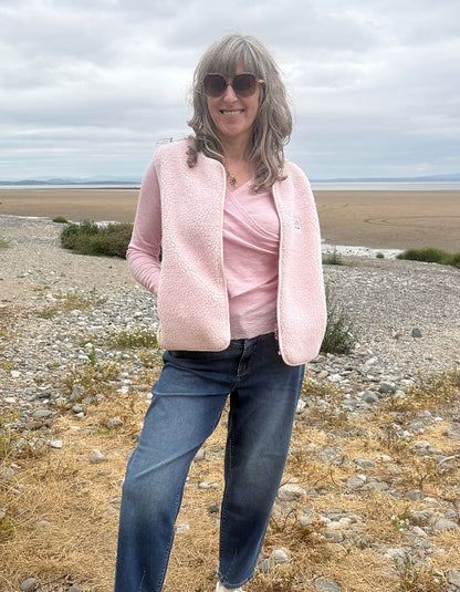 A woman wearing a light pink sherpa zip-up vest over a matching pink top, blue jeans, and sunglasses, standing outdoors on a rocky, dry beach landscape under an overcast sky.