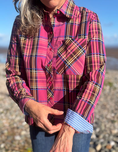 A woman wearing a long-sleeve plaid button-up shirt in pink, red, and brown tones, with blue jeans, photographed outdoors on a rocky beach with water and sky in the background.