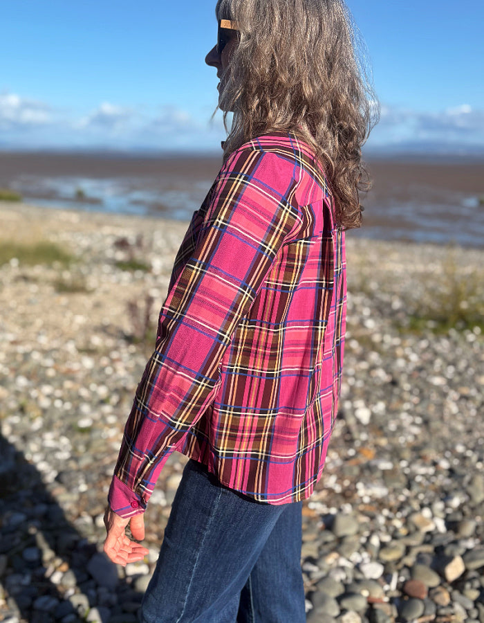 A person standing outdoors on a pebble beach wearing a bright pink plaid button-up shirt with blue jeans, photographed from the side in natural daylight.