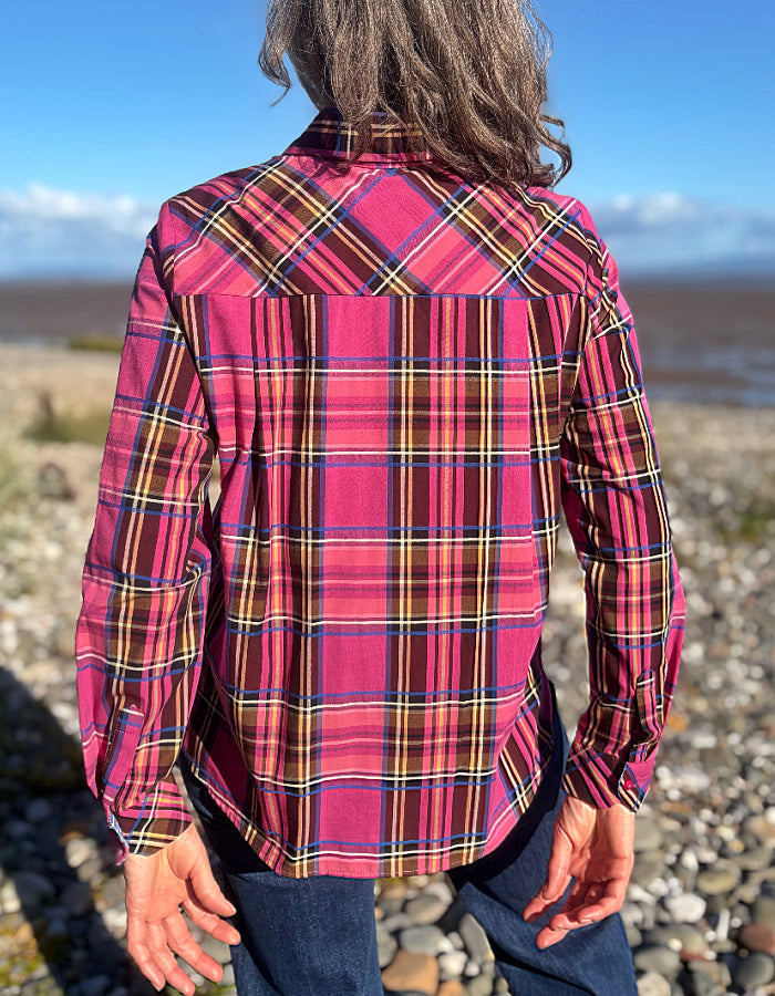 A person standing outdoors on a rocky beach, viewed from the back, wearing a long-sleeve plaid button-up shirt in bright pink with yellow, blue, and black stripes, paired with dark blue jeans.