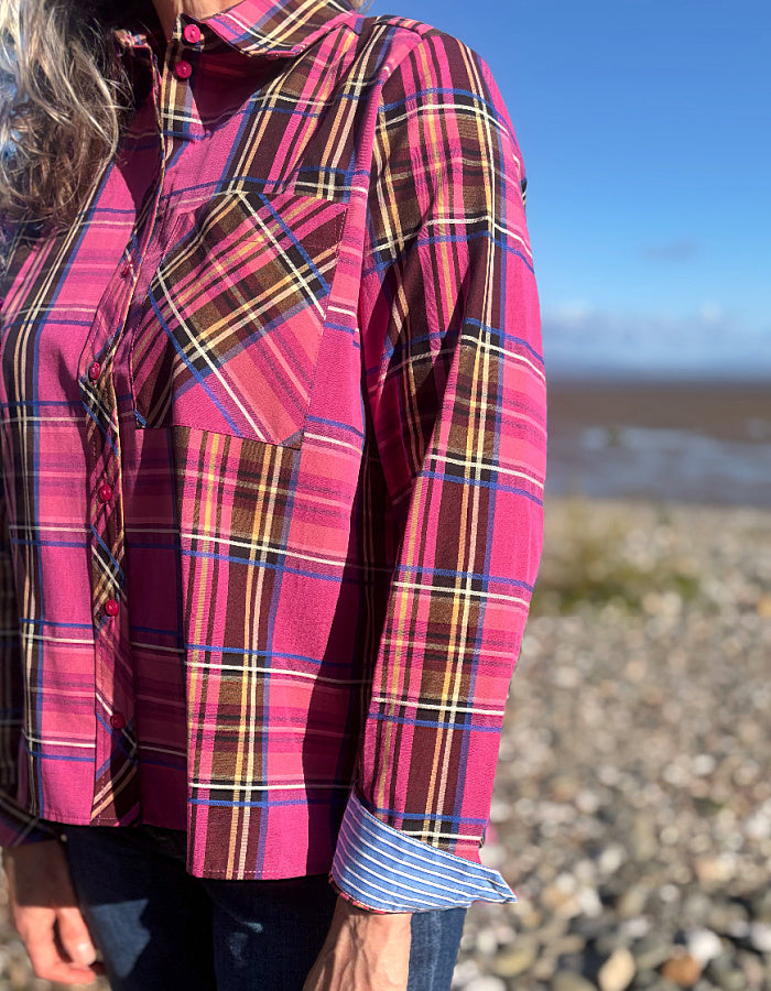 A close-up of a person wearing a bright pink plaid button-up shirt with a chest pocket, standing outdoors on a rocky beach with water and blue sky in the background.