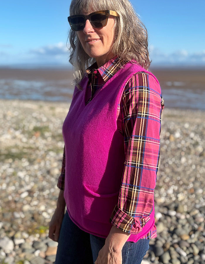 A woman wearing a bright pink fleece vest over a long-sleeve plaid shirt, standing on a rocky beach with water and sky in the background.
