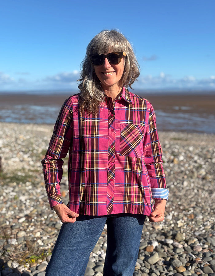 A woman wearing a pink plaid button-up shirt and blue jeans stands on a rocky beach with water and a blue sky in the background.