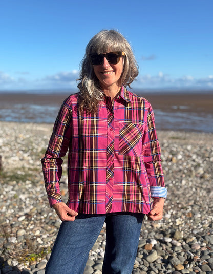 A woman wearing a pink plaid button-up shirt and blue jeans stands on a rocky beach with water and a blue sky in the background.