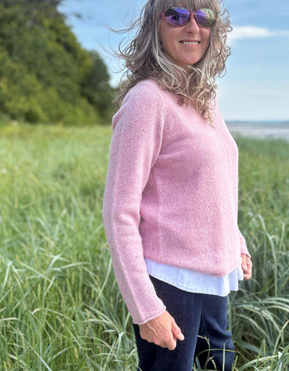Woman in a pink wool sweater standing in tall grass by a coastal area