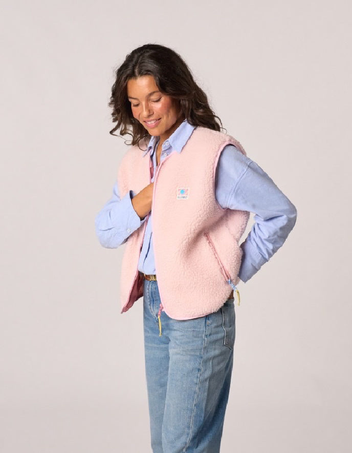 A woman wearing a light pink fleece vest over a light blue button-down shirt and blue jeans, standing against a solid light gray studio background.