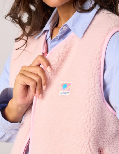 A close-up of a person wearing a light pink fleece vest over a light blue collared shirt. The vest has a small square HellyBelt logo patch on the left chest and pink piping along the edges.