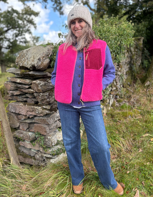 Person wearing a bright pink vest and blue jeans standing in a natural setting with a stone wall and greenery.