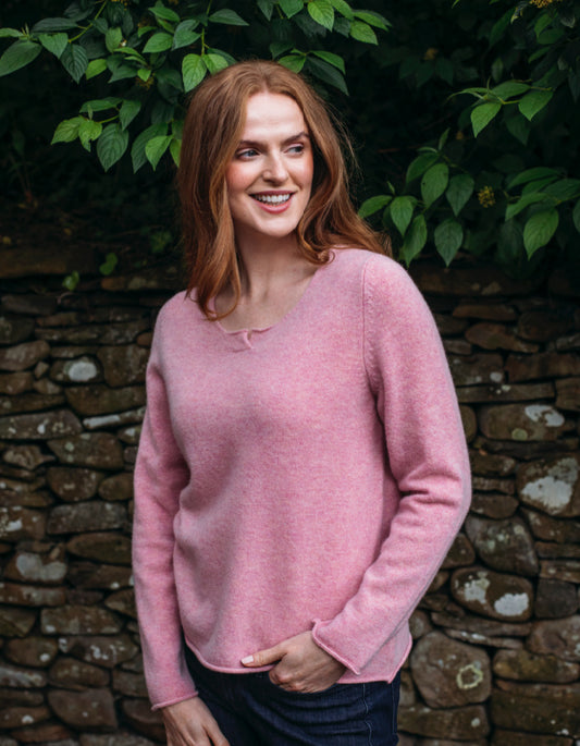 Woman wearing a pink sweater standing in front of a stone wall with greenery.