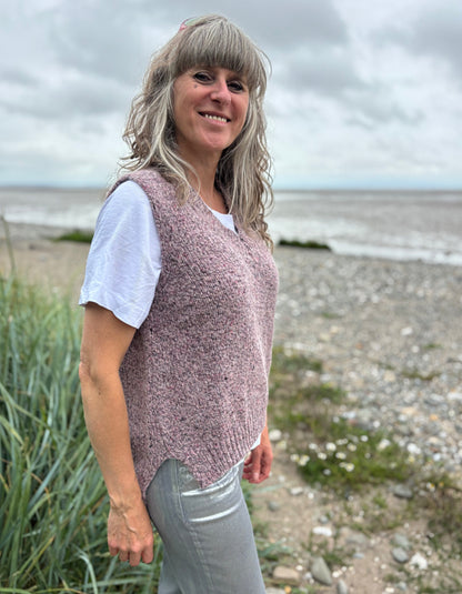 Woman standing on a beach wearing a pink knitted vest and white shirt.