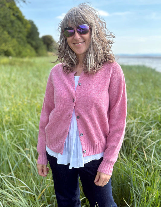 Woman wearing a short pink wool cardigan in a grassy field with a scenic background. 