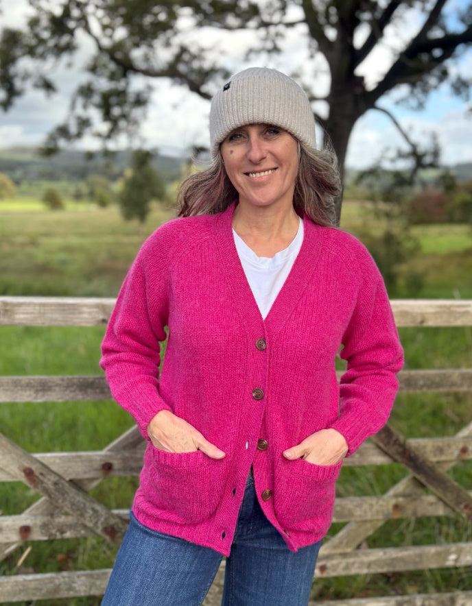 Woman wearing a pink cardigan and gray beanie standing in front of a wooden fence with a scenic background.