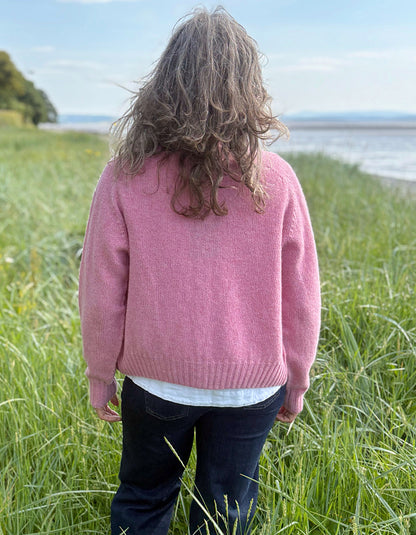 Woman wearing a short pink wool cardigan in a grassy field with a scenic background. 