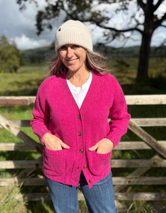 Woman wearing a pink cardigan, white beanie, and blue jeans standing outdoors with a wooden fence and trees in the background.