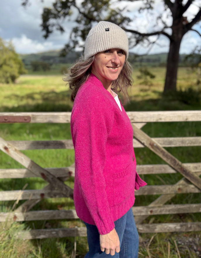 Woman wearing a pink sweater and white beanie standing in front of a wooden gate with a scenic background.