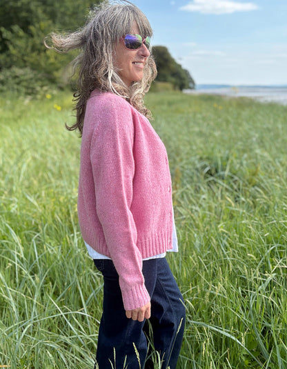 Woman wearing a short pink wool cardigan in a grassy field with a scenic background. 