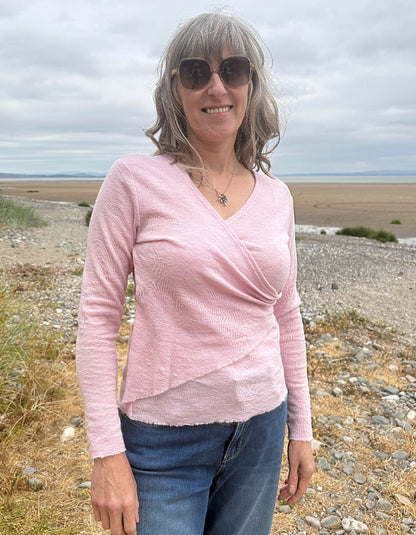 A woman with gray hair and sunglasses wearing a light pink textured wrap-style long-sleeve top and blue jeans, standing outdoors on a rocky beach or shoreline under an overcast sky.