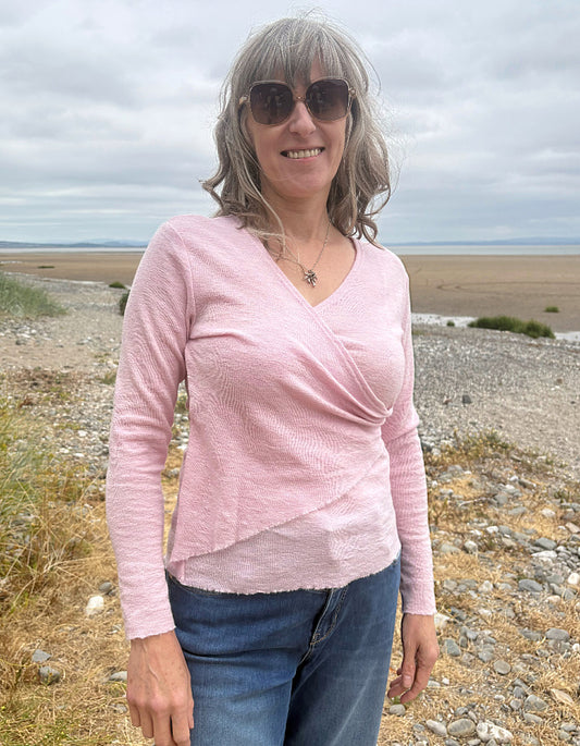 A woman with gray hair and sunglasses wearing a light pink textured wrap-style long-sleeve top and blue jeans, standing outdoors on a rocky beach or shoreline under an overcast sky.