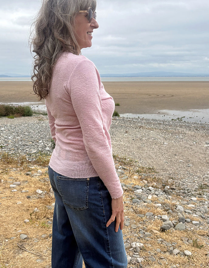 A woman with long wavy gray hair and sunglasses, wearing a light pink ribbed long-sleeve top and dark blue jeans, standing on a rocky beach with water and a cloudy sky in the background.