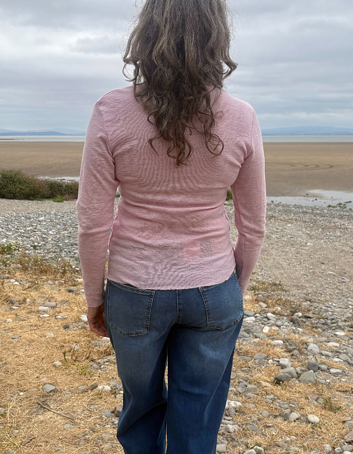 A woman viewed from behind wearing a light pink long-sleeve textured knit top and blue jeans, standing on a rocky beach with a cloudy sky in the background.