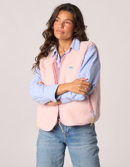 A woman wearing a light pink sherpa fleece vest over a light blue button-down shirt and blue jeans, standing against a light gray studio background.