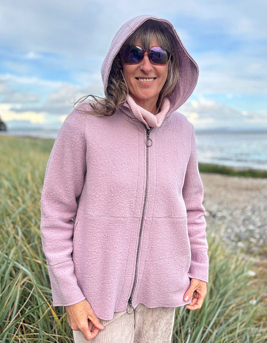 Person wearing a pink boiled wool hoodie jacket on a beach with grass and water in the background