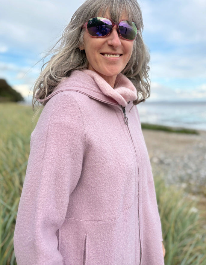 Person wearing a pink boiled wool hoodie jacket on a beach with grass and water in the background