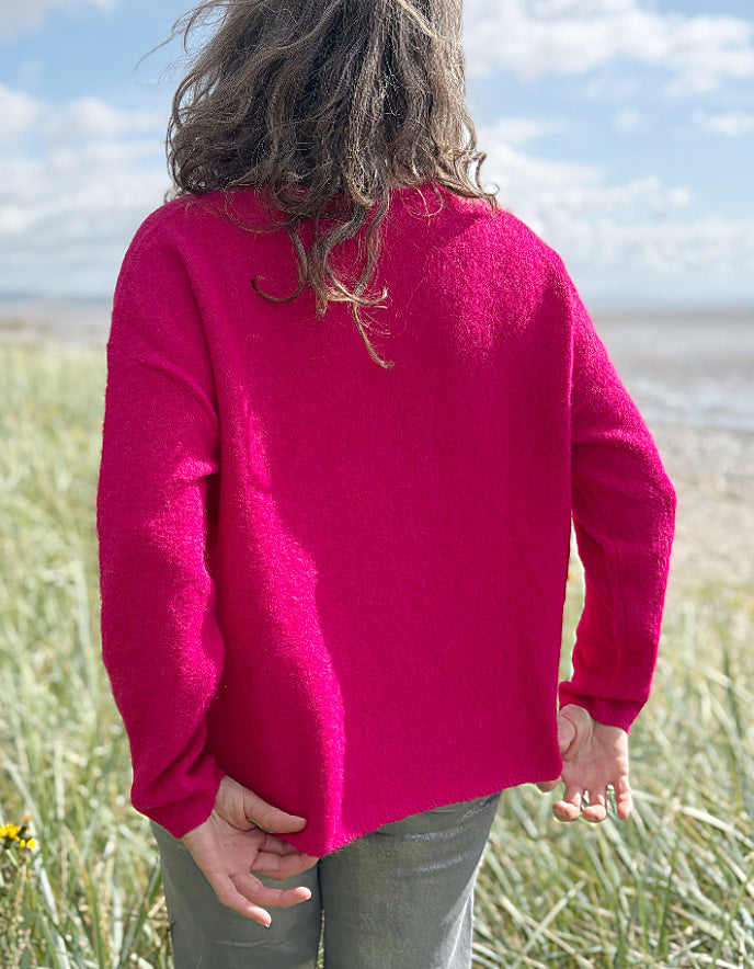 Woman in a pink v neck sweater and sunglasses standing in grass near a beach.