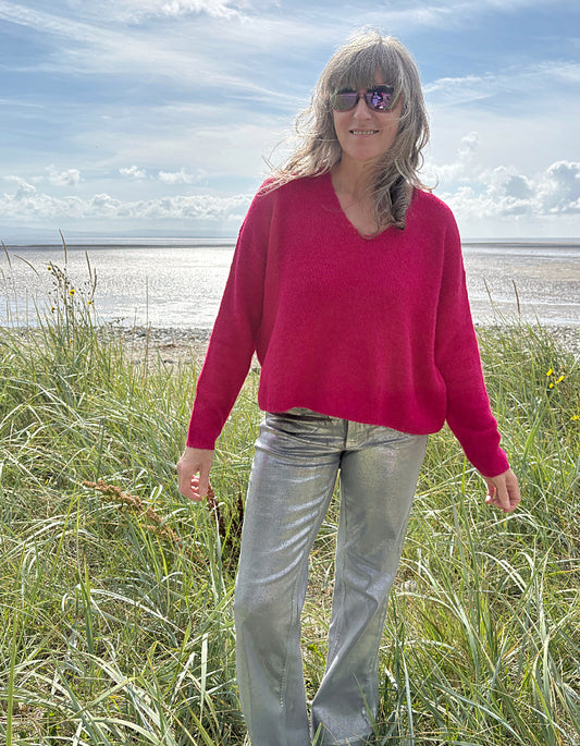 Woman in a pink v neck sweater and sunglasses standing in grass near a beach.
