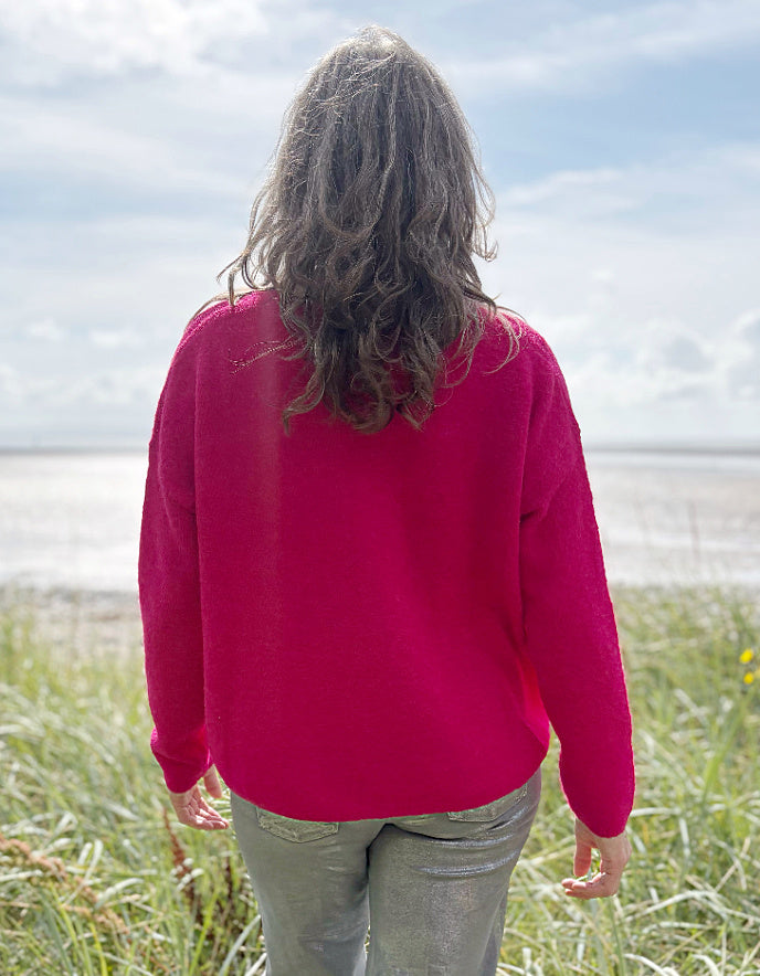 Woman in a pink v neck sweater and sunglasses standing in grass near a beach.