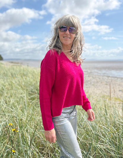Woman in a pink v neck sweater and sunglasses standing in grass near a beach.