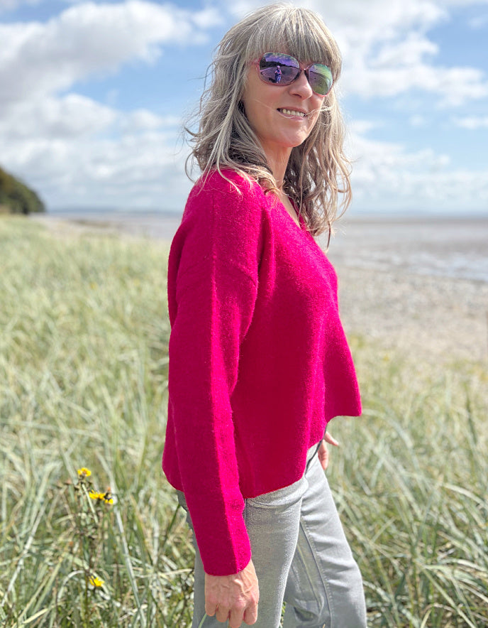 Woman in a pink v neck sweater and sunglasses standing in grass near a beach.
