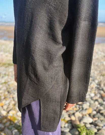 A close-up of a person outdoors wearing a dark charcoal gray textured long-sleeve top or cardigan over purple striped pants, standing on a rocky beach.