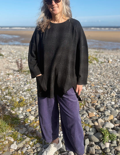 A woman wearing a black oversized knit sweater and purple corduroy trousers stands on a rocky beach with the ocean and sand visible in the background.