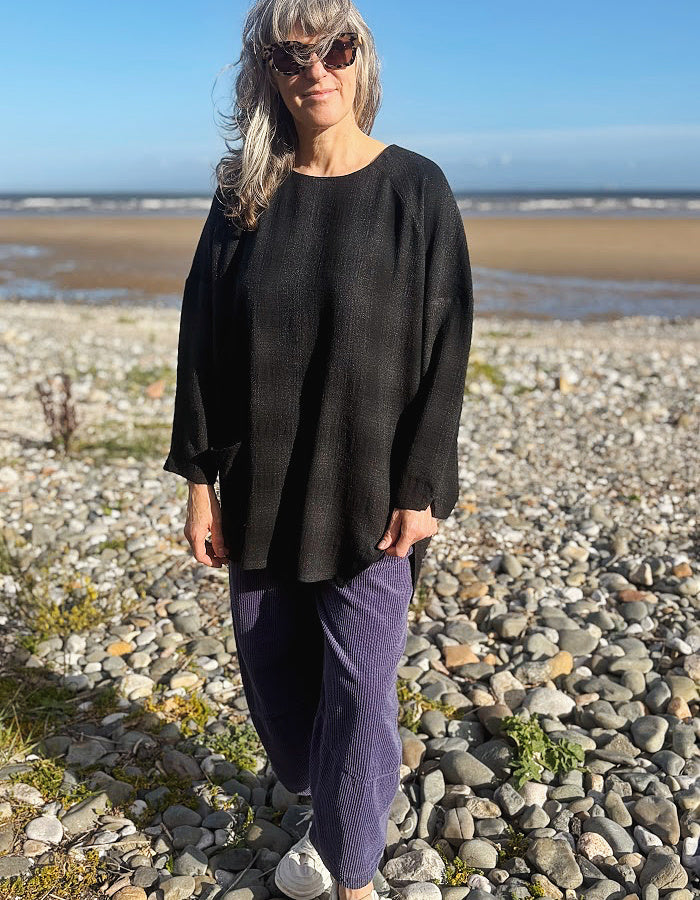 A woman wearing a loose black long-sleeve top and purple pants stands on a rocky beach with the ocean and blue sky in the background.