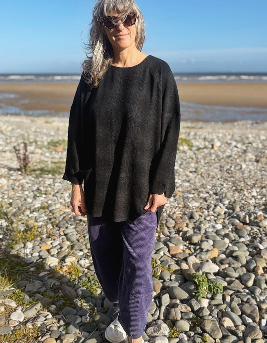 A woman wearing a loose black long-sleeve top and purple pants stands on a rocky beach with the ocean and blue sky in the background.