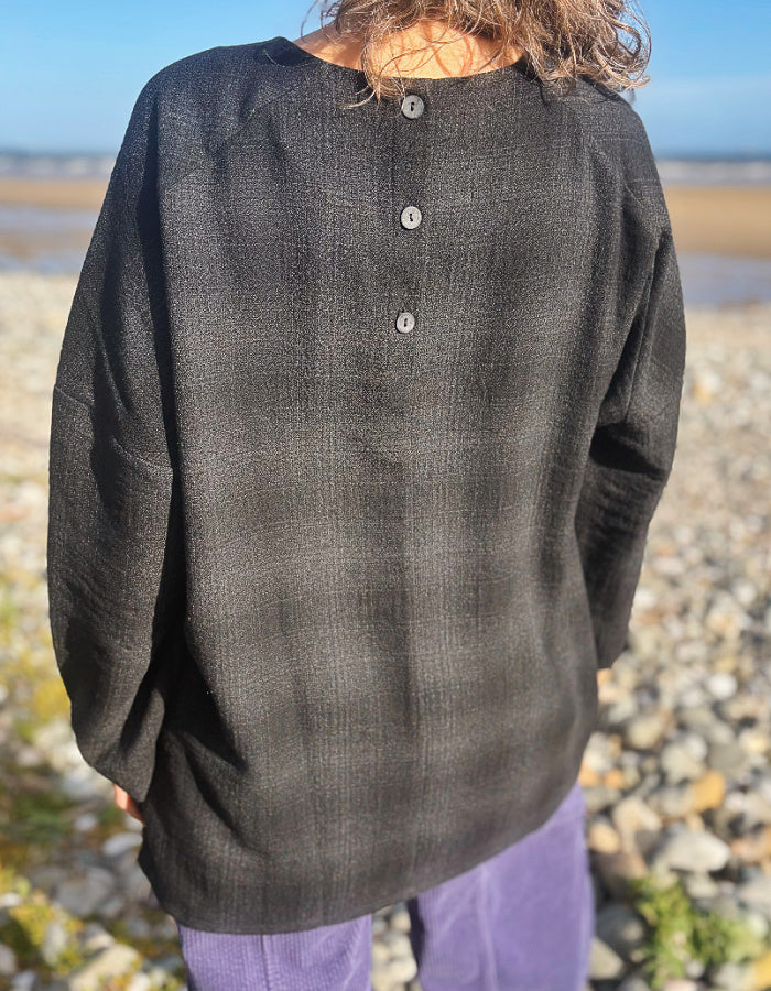 A person standing on a rocky beach wearing a loose, dark charcoal gray textured long-sleeve top with three buttons at the back neckline, paired with purple pants.