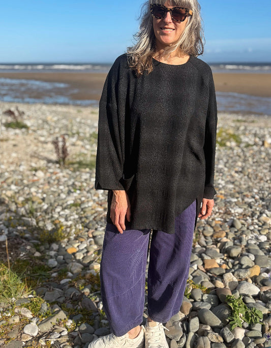 A woman wearing a black textured knit long-sleeve top and purple corduroy pants stands on a rocky beach.