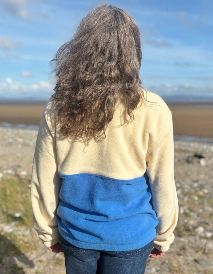 Person wearing a beige and blue jacket standing on a beach with a scenic view.