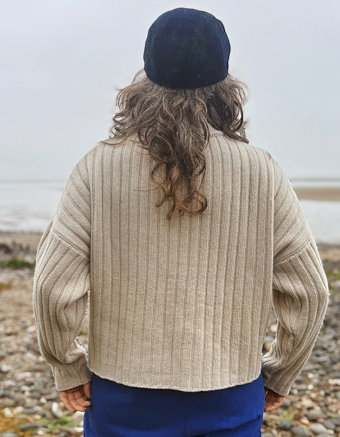 A person viewed from behind wearing a beige ribbed knit sweater, a dark navy beret, and blue pants, standing on a pebble beach with water in the background.