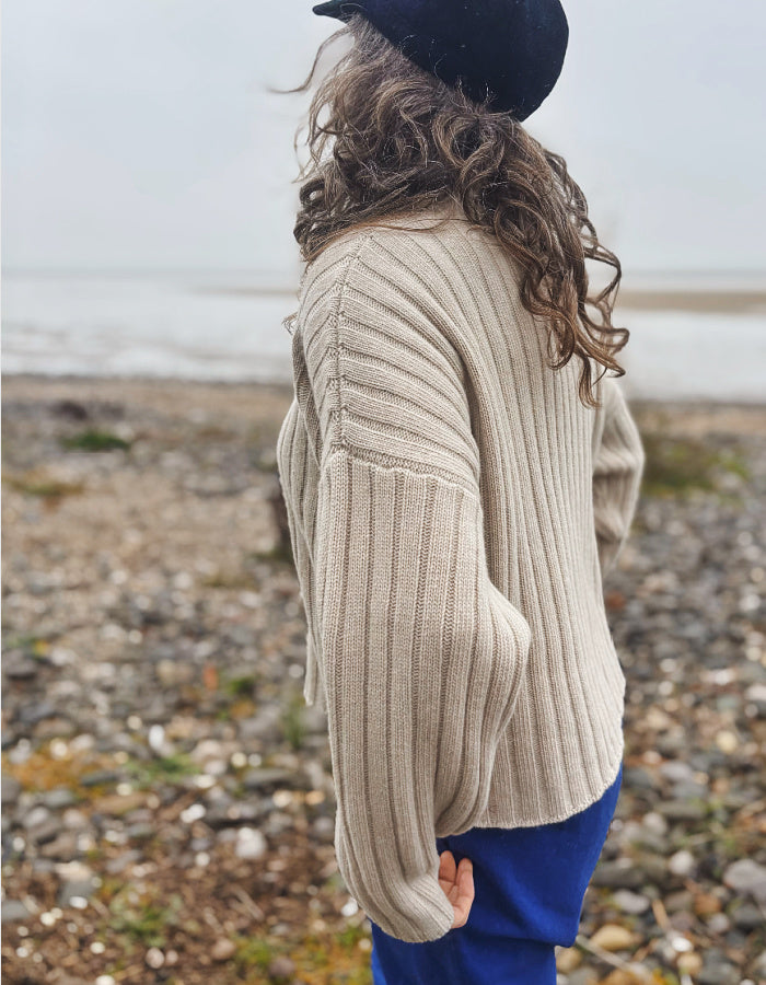 A person standing on a pebble beach wearing a cream ribbed knit sweater, blue pants, and a black hat, photographed from the back.