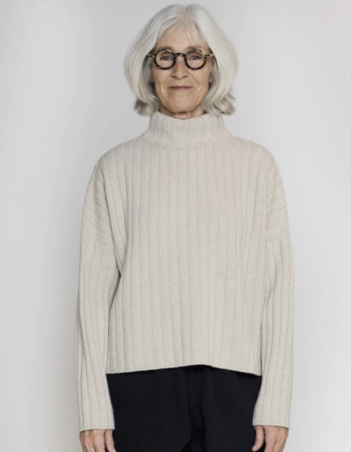 An older woman with short gray hair and glasses wearing a cream ribbed knit mock-neck sweater and black pants, standing against a light gray studio background.