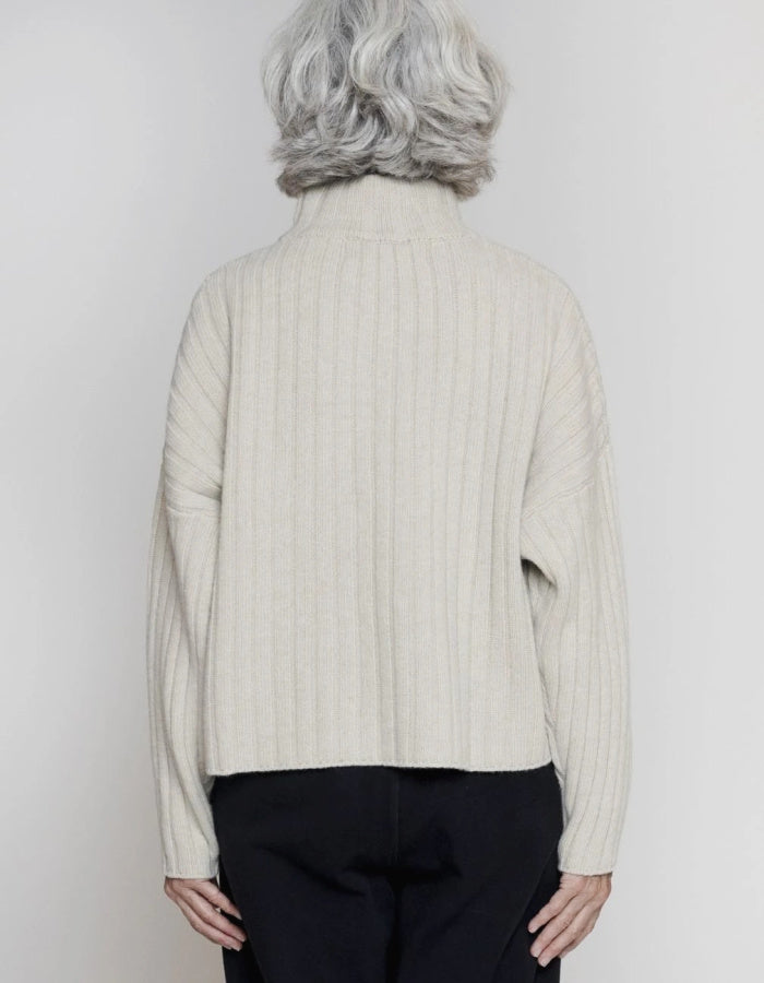 A person shown from the back wearing a cream ribbed knit turtleneck sweater and black pants against a plain light gray studio background.