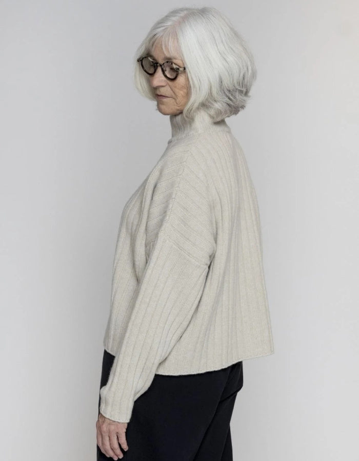 An older woman with short white hair and glasses, shown in profile wearing a ribbed knit cropped sweater in a light cream or off-white color, paired with black pants, against a plain light gray studio background.